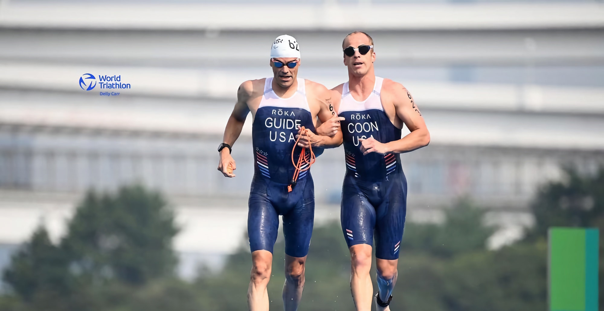 Two athletes running out of the water during the Paralympic Games in Tokyo. They are both wearing blue triathlon suits with USA written on them. The man on the right of the picture has Guide written on his suit and the man on the left has the name Coon written on his. They are both wearing swim caps and goggles.